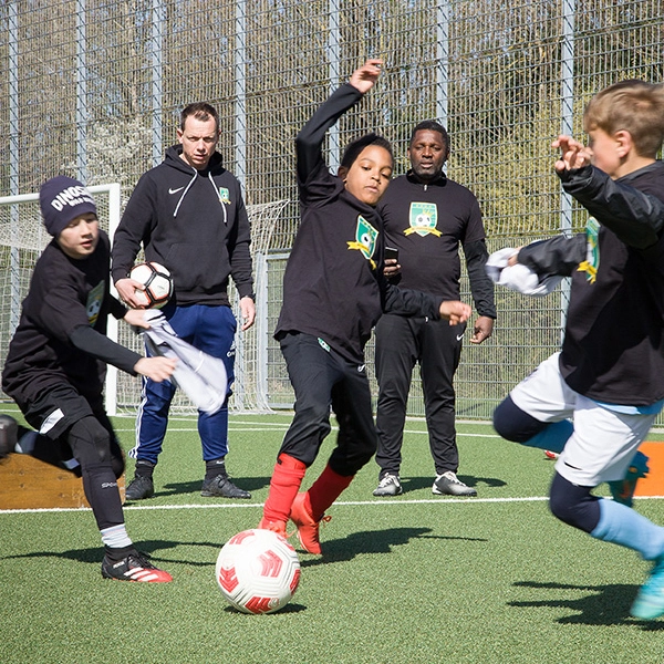 Kinder trainieren Fußball auf dem Platz unter Anleitung von zwei Trainern der ABC-Fußballschule, die aufmerksam das Training beobachten. Ein Kind führt den Ball, während andere Spieler in Bewegung sind.