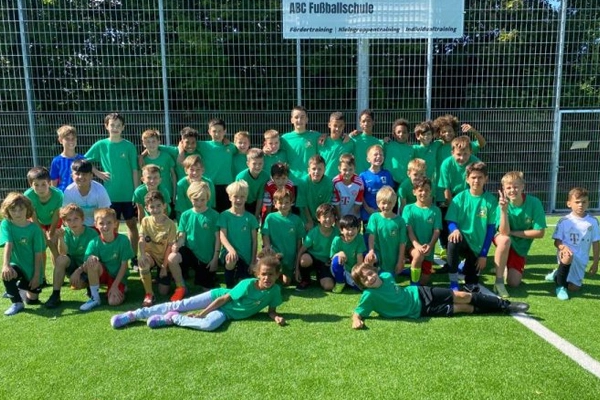 Eine große Gruppe von Kindern posiert auf einem Fußballfeld für ein Foto während eines Feriencamps der ABC Fußballschule. Alle tragen grüne T-Shirts und lächeln in die Kamera, hinter ihnen ist ein Schild der Fußballschule zu sehen.