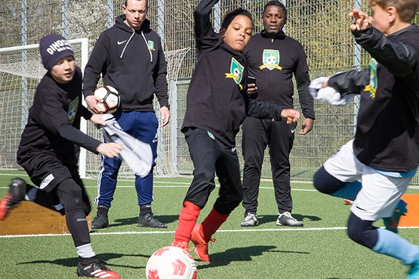Junge Spieler der ABC Fußballschule beim Einzeltraining auf dem Fußballplatz, zwei Trainer im Hintergrund beobachten die Szene. Ein Spieler in roten Stutzen dribbelt den Ball, während zwei andere Spieler versuchen, ihn zu stoppen.
