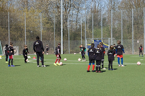 Ein Jugendtraining der ABC Fußballschule auf einem Fußballfeld, bei dem mehrere Kinder in schwarzen Trainingsanzügen Übungen mit dem Ball durchführen. Trainer und Spieler sind über das ganze Feld verteilt.