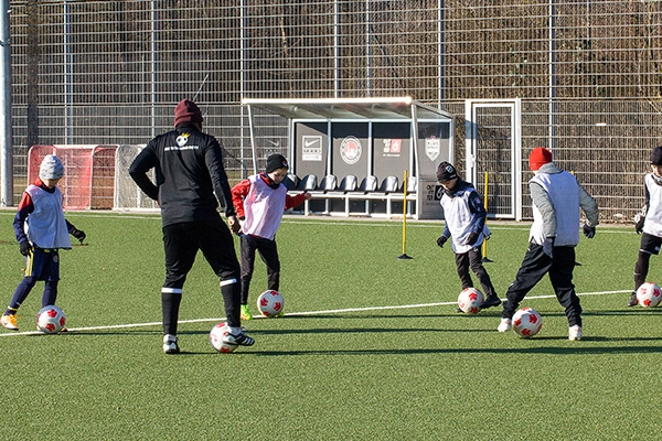 Ein Trainer der ABC Fußballschule leitet ein Training auf dem Fußballplatz, während mehrere Kinder mit Mützen und Westen Übungen mit dem Ball machen. Im Hintergrund befindet sich eine Ersatzbank.