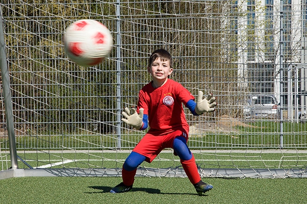 Ein junger Torwart der ABC Fußballschule im roten Trikot trainiert auf dem Fußballplatz. Er steht vor dem Tor und bereitet sich darauf vor, einen heranfliegenden Ball zu fangen.