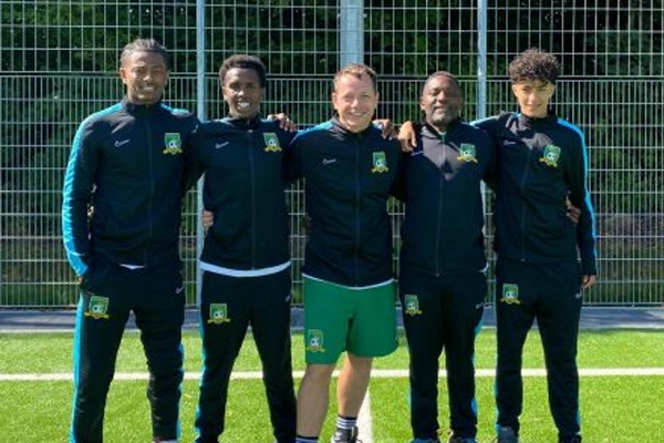 Das Trainerteam der ABC Fußballschule steht auf einem Fußballfeld vor einem Metallzaun, alle in Trainingsanzügen des Vereins gekleidet. Sie lächeln und legen die Arme umeinander.